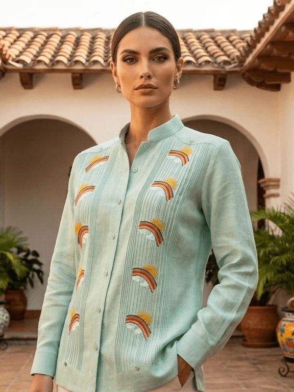 Camisa guayabera de manga larga en azul turquesa pálido con bordados de soles, nubes y arcoíris coloridos, presentada en un maniquí sobre un fondo artístico.
