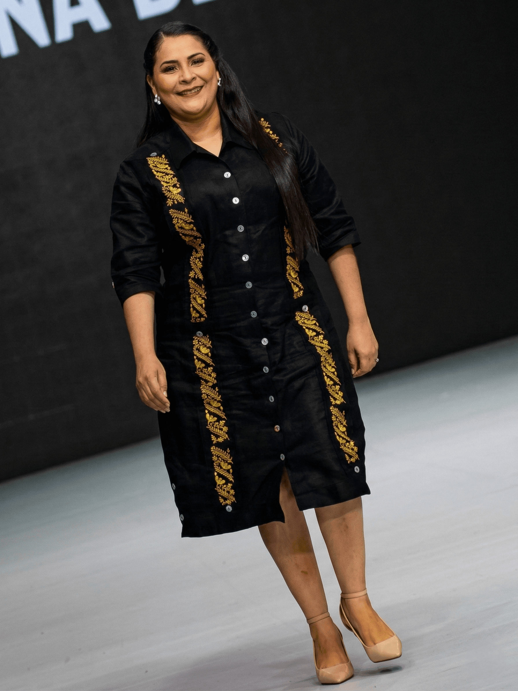 Vestido tipo camisero de color negro intenso con dos franjas verticales de bordados florales en hilo dorado brillante. El diseño presenta botones frontales blancos, cuello estructurado y mangas 3/4. Se aprecian detalles de botones en las aberturas laterales inferiores sobre un fondo gris neutro