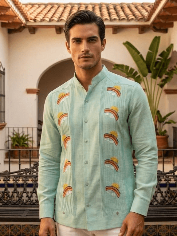 Camisa guayabera de manga larga en azul turquesa pálido con bordados de soles, nubes y arcoíris coloridos, presentada en un maniquí sobre un fondo artístico.